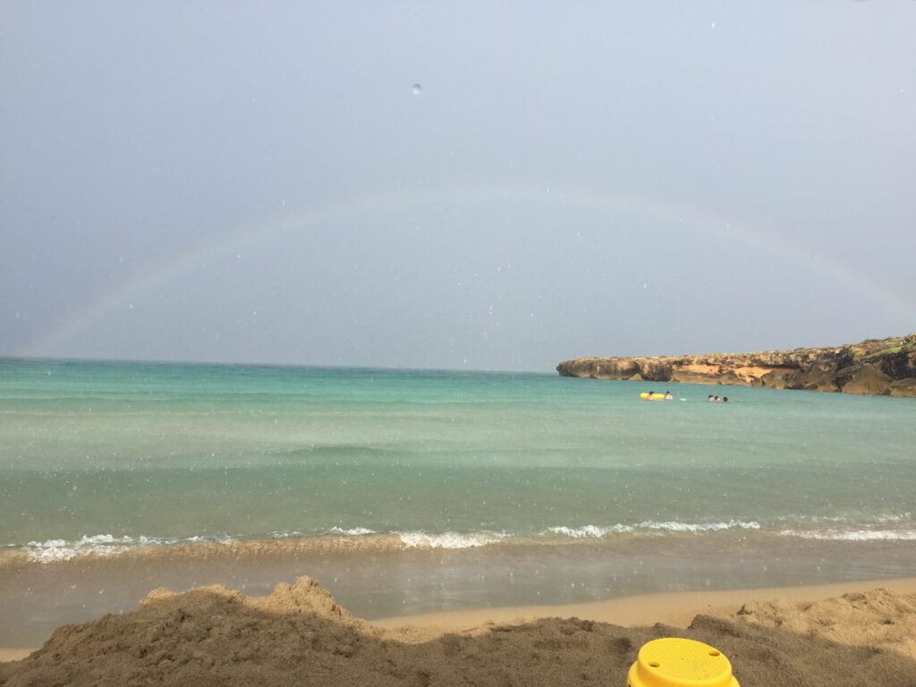 Spiaggia di Calamosche vista dalla battigia, con mare calmo e un arcobaleno visibile sopra l’orizzonte.