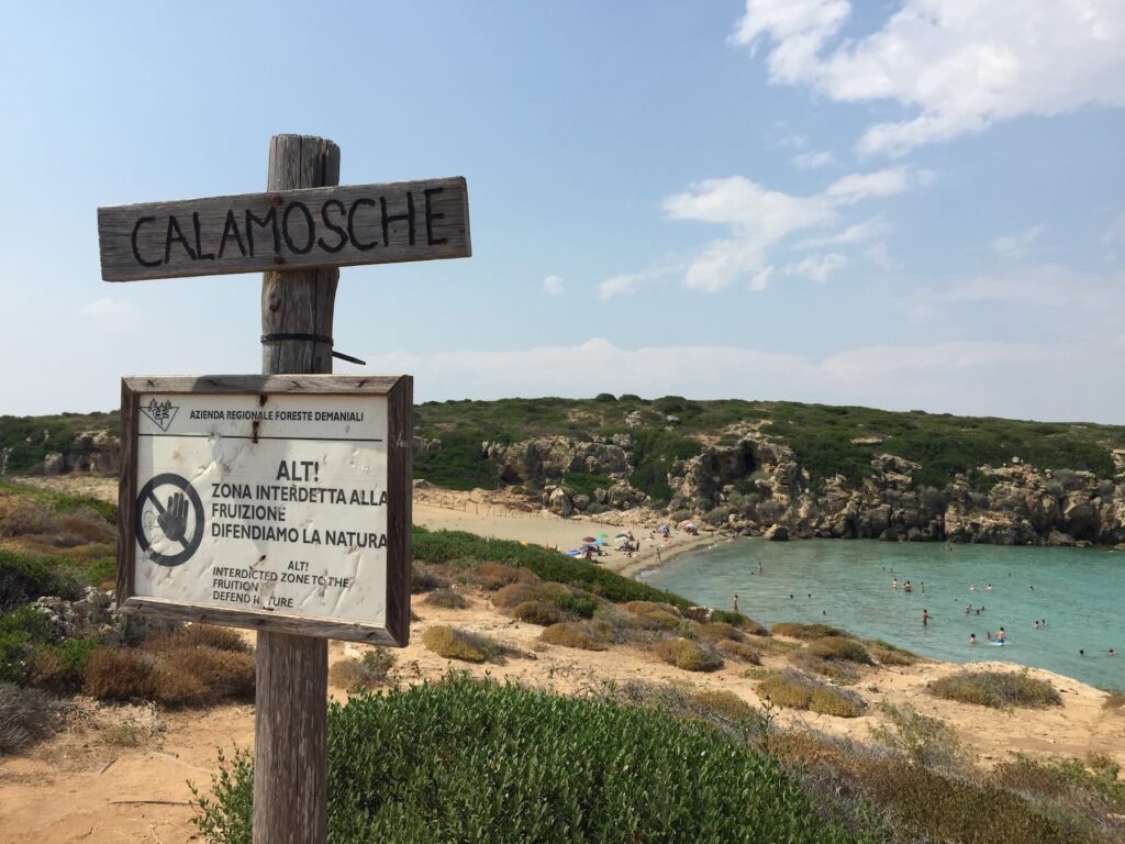 Cartello in legno con la scritta “Calamosche” e segnalazione di area interdetta per protezione ambientale, con vista sulla spiaggia sullo sfondo.