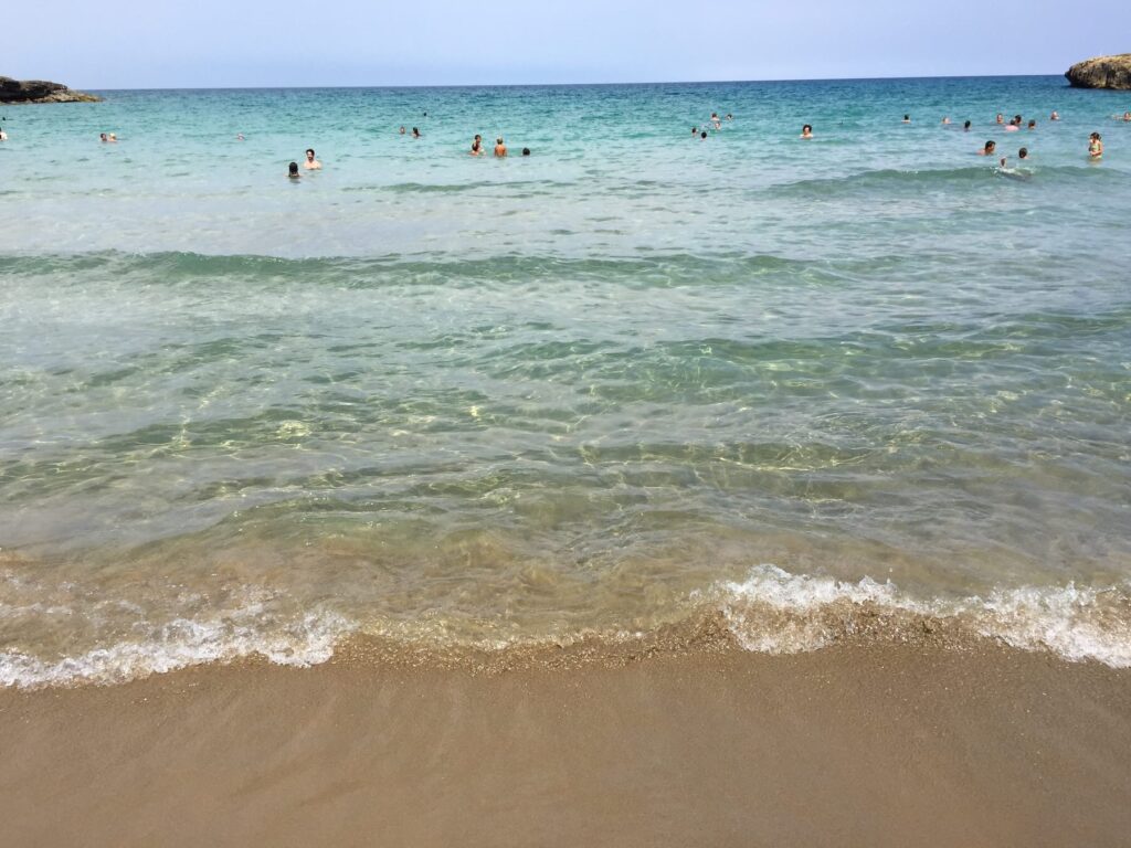 Primo piano del mare cristallino e della sabbia dorata a Calamosche, con alcune persone che nuotano in acqua.