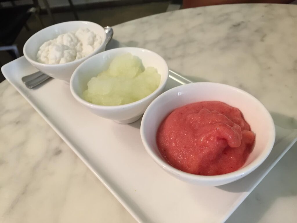 Tre ciotoline di granita servite su un vassoio: da sinistra a destra, gusto mandorla, limone e fragola, fotografate su un tavolo di marmo in un bar di Noto.