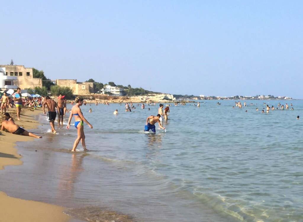 Persone che si godono il mare limpido e la sabbia dorata a Marina di Noto, una spiaggia in una giornata d’estate, con edifici bassi sullo sfondo e cielo sereno.