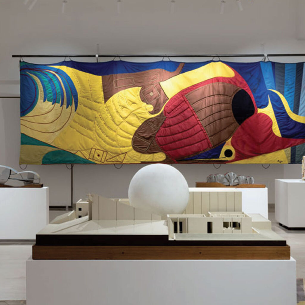 Interno del MAC Gibellina con modello architettonico bianco in primo piano e grande arazzo colorato giallo e blu sullo sfondo, esempio delle opere d'arte contemporanea esposte nella capitale italiana dell'arte 2026.