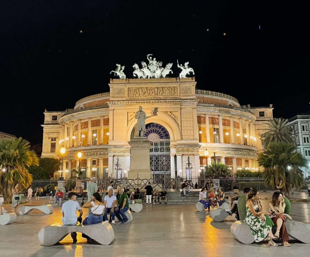 Vista notturna della facciata del Teatro Politeama Garibaldi di Palermo illuminata da luci calde. In primo piano, gruppi di persone sedute su panchine moderne in pietra nella piazza antistante. Al centro, l'imponente arco di trionfo del teatro con la statua equestre in bronzo della Quadriga sulla sommità. L'architettura presenta colonne classiche e decorazioni dettagliate, stagliandosi contro il cielo scuro.