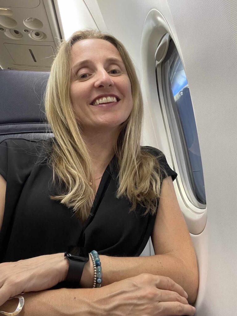 Donna sorridente seduta vicino al finestrino di un aereo durante un volo verso la Sicilia.