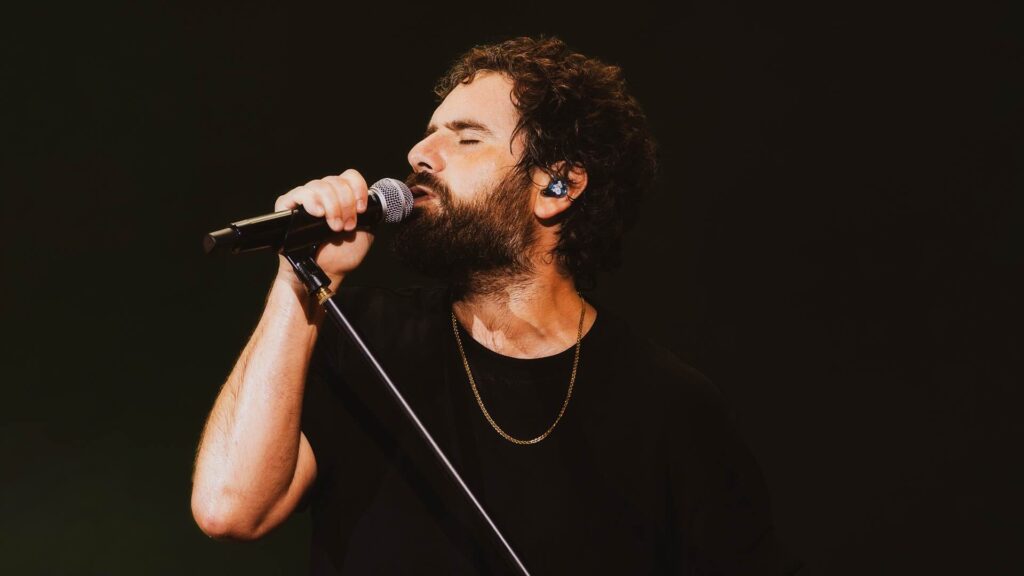 Tommaso Paradiso canta sul palco durante un concerto, con microfono in mano e sfondo scuro. Foto divulgazione.