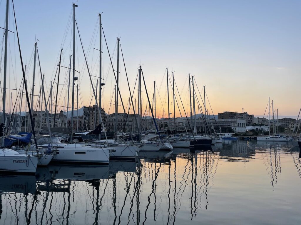 Caparezza a Palermo 2026: biglietti, prezzi e consigli per il Velodromo Borsellino 3 Barche a vela ormeggiate alla Cala di Palermo al tramonto, con il profilo della città sullo sfondo e i riflessi degli alberi sull’acqua.